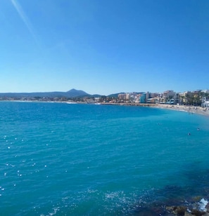 A coastal view with a clear blue sea, a sandy beach, and a cityscape in the background.