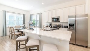 A modern kitchen with white cabinets, stainless steel appliances, a central island with stools, and a dining area with chairs.
