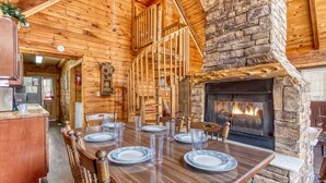 A wooden dining area with a stone fireplace, wooden ceiling beams, and a view of the kitchen.