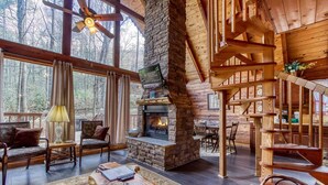 A cozy cabin interior with a stone fireplace, wooden furniture, and large windows offering a forest view.