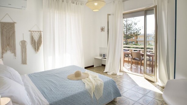 A bedroom with a bed, a bedside table, a hanging macrame wall decor, a window with curtains, and a view of the outside.