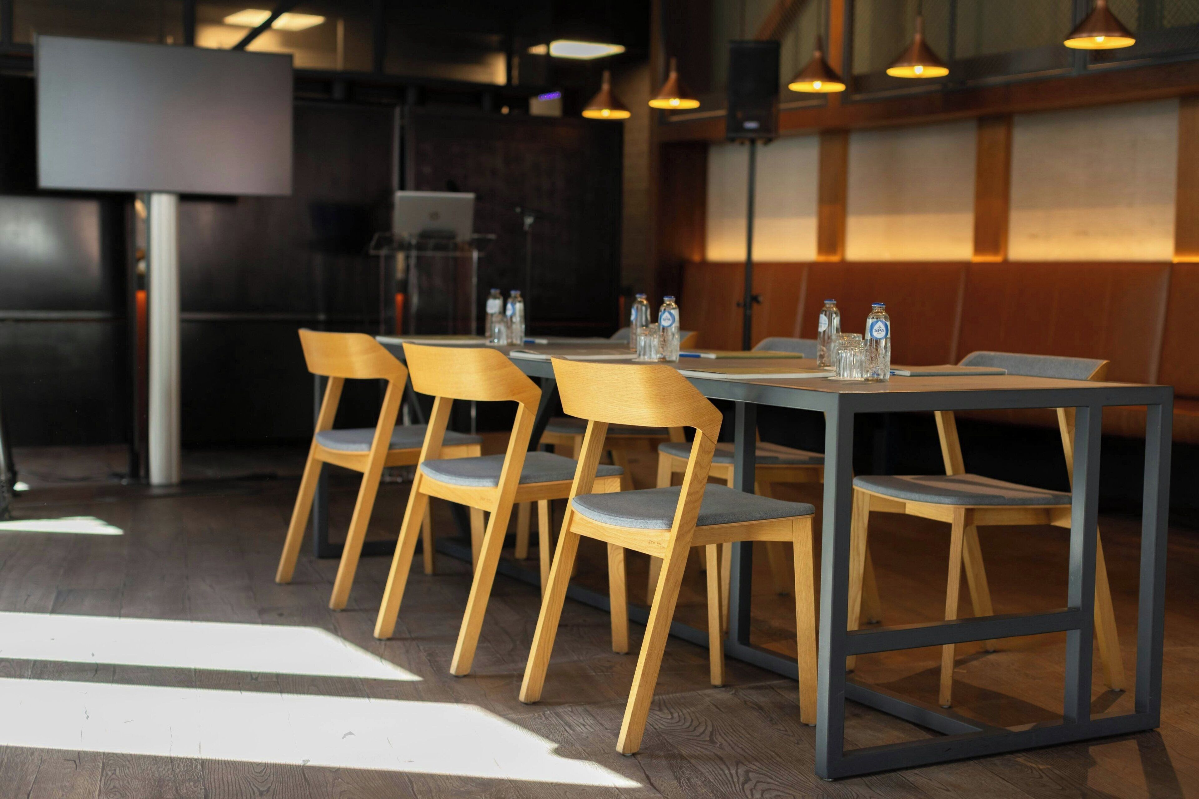 Un restaurante con sillas y mesas de madera, una pantalla grande e iluminación colgante.