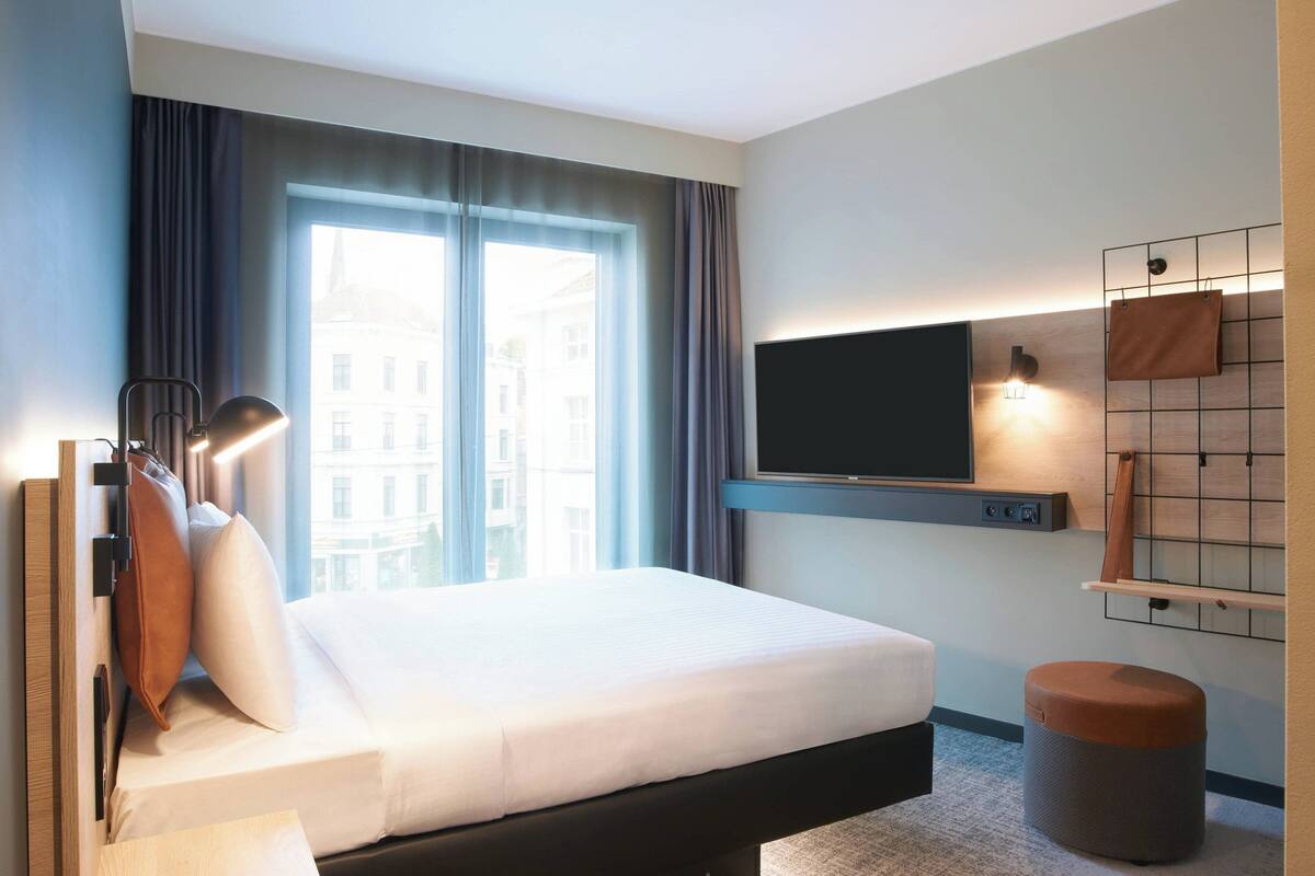 A modern hotel room with a large bed, a TV mounted on the wall, a small stool, and a view of the city through the window.