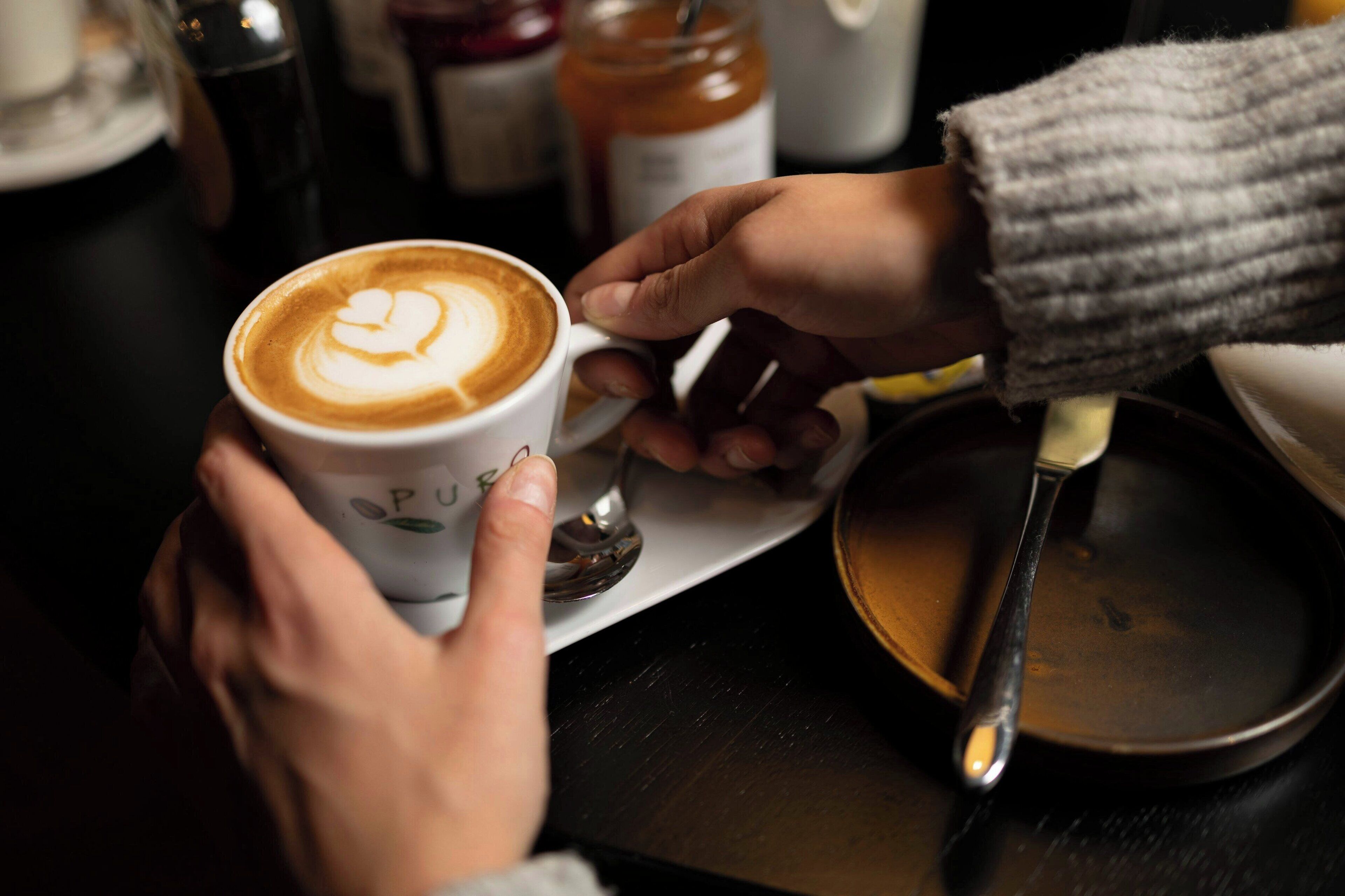 A person holding a cup of coffee with a heart design on the foam, a spoon, and a saucer on a table.