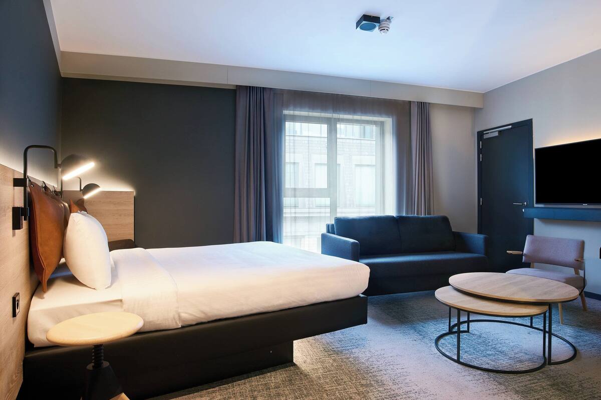 A modern hotel room with a bed, a small round table, a sofa, and a TV.