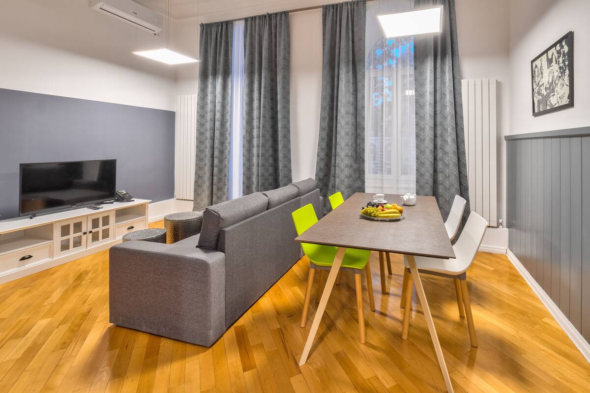 A modern living room with a grey sofa, a dining table with chairs, and a flat-screen TV.