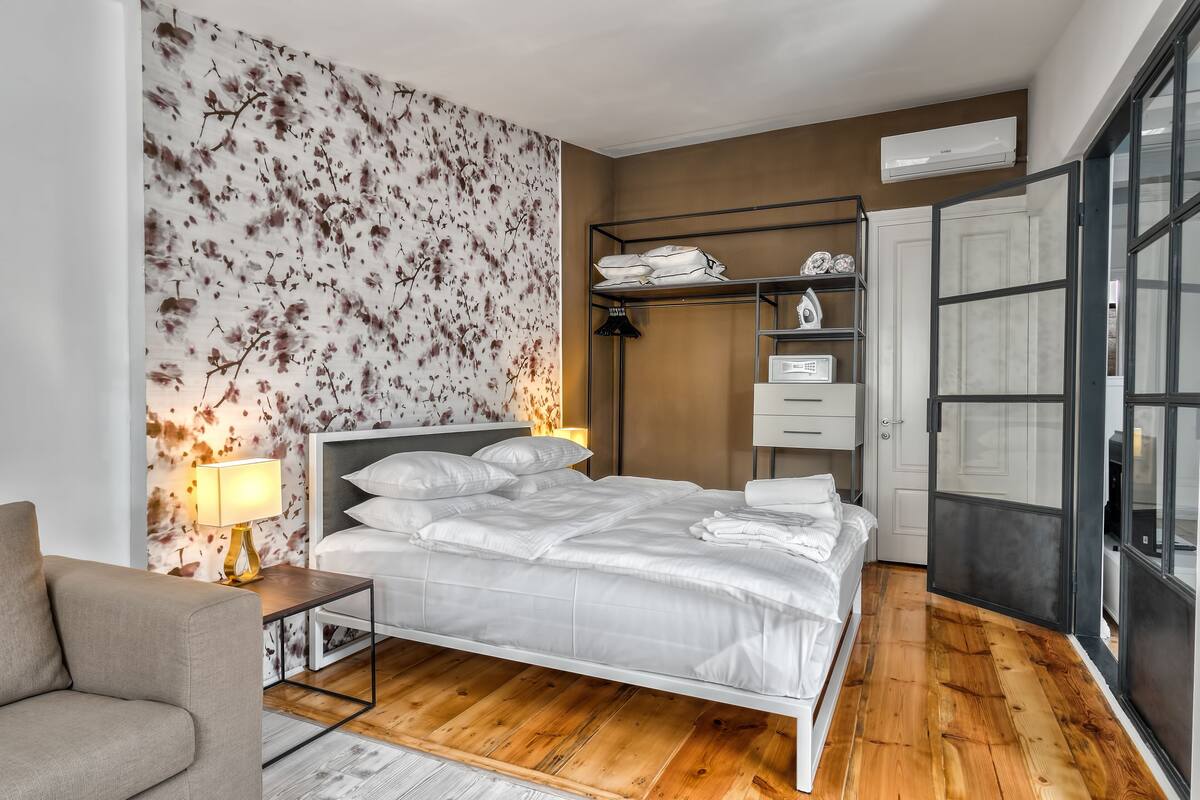 A modern bedroom with a large bed, a bedside table, a lamp, a wardrobe, and a view through a glass door.