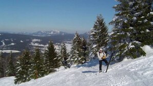 Uma pessoa está em uma encosta coberta de neve, com vista para montanhas e árvores ao longe.