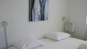 A hotel room with a bed, white pillows, a wall-mounted artwork, and a small white cabinet. - Plesners Anneks (Skagen)