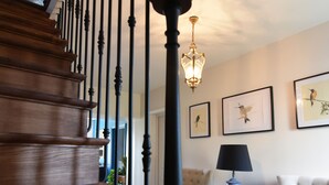 Una escalera de madera con barandillas negras que conduce a una plataforma. Hay cuadros enmarcados en la pared y una lámpara sobre una mesita.