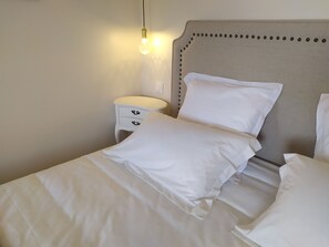 Una cama bien hecha con ropa de cama blanca, una mesita de noche con cajones y una lámpara fijada a la pared.