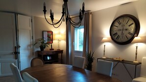 Comedor con una mesa redonda de madera, sillas blancas, una lámpara de araña, un reloj de pared y una lámpara.