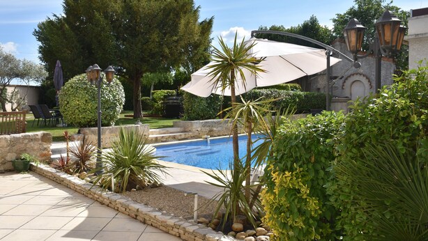 Una piscina rodeada por un jardín ajardinado con diversas plantas y una pérgola con toldo blanco.