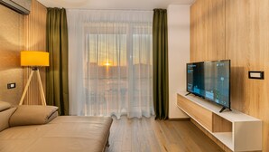 A modern hotel room with a bed, a TV on a stand, a lamp, and a view of the outdoors through sheer curtains. - Promenada Studios (Brasov)