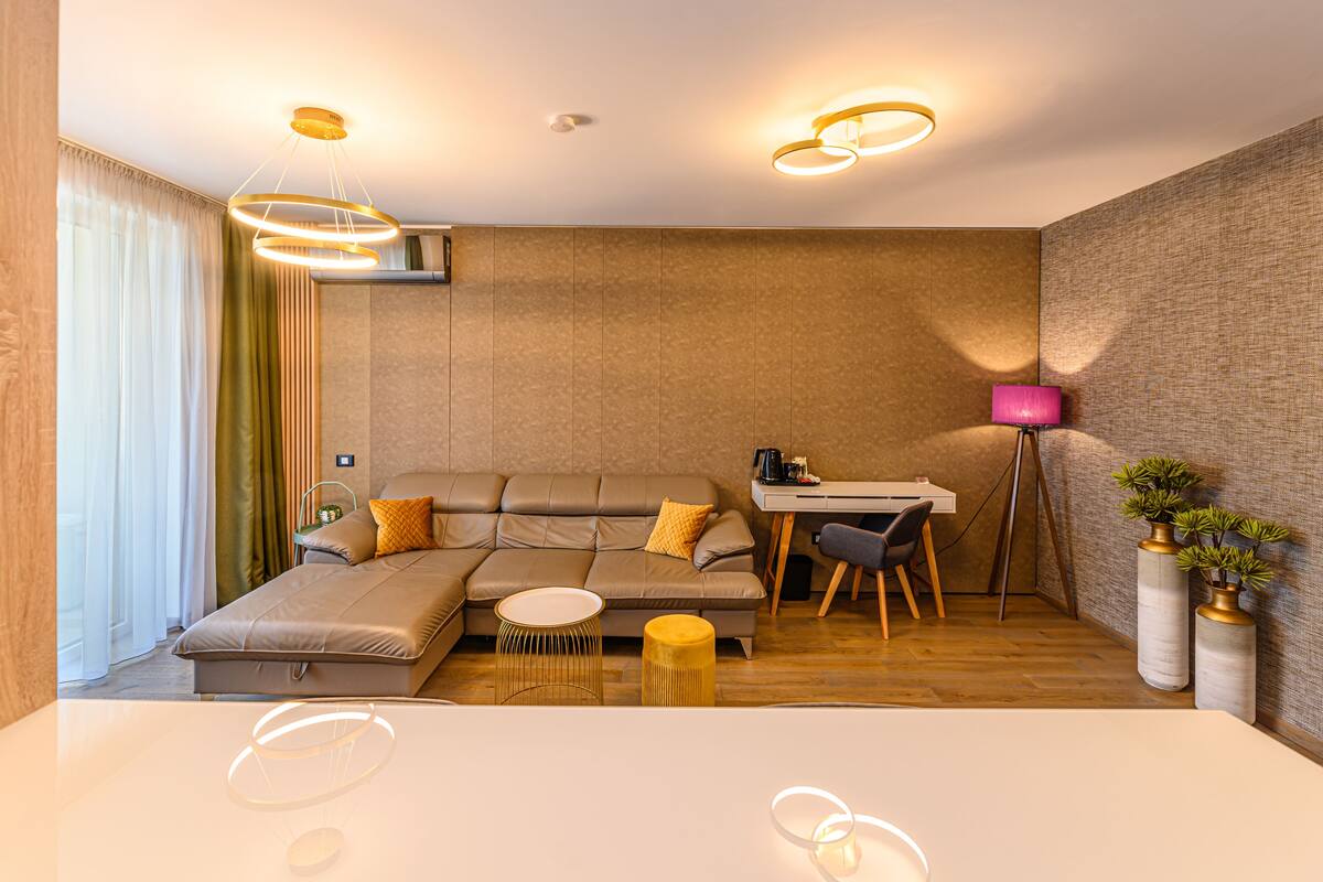 Quarto de hotel moderno com sofá em forma de L, uma mesa redonda pequena, escrivaninha e uma cadeira.