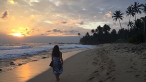 A beach scene with a person walking along the shore, palm trees in the background, and the sun setting over the ocean.