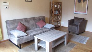 Uma sala de estar com um sofá cinzento, uma mesa de centro de madeira, uma poltrona marrom e uma estante de livros.