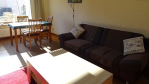 Uma sala de estar aconchegante com uma mesa de madeira, duas cadeiras, um sofá com almofadas estampadas, um quadro na parede e uma janela com cortinas.
