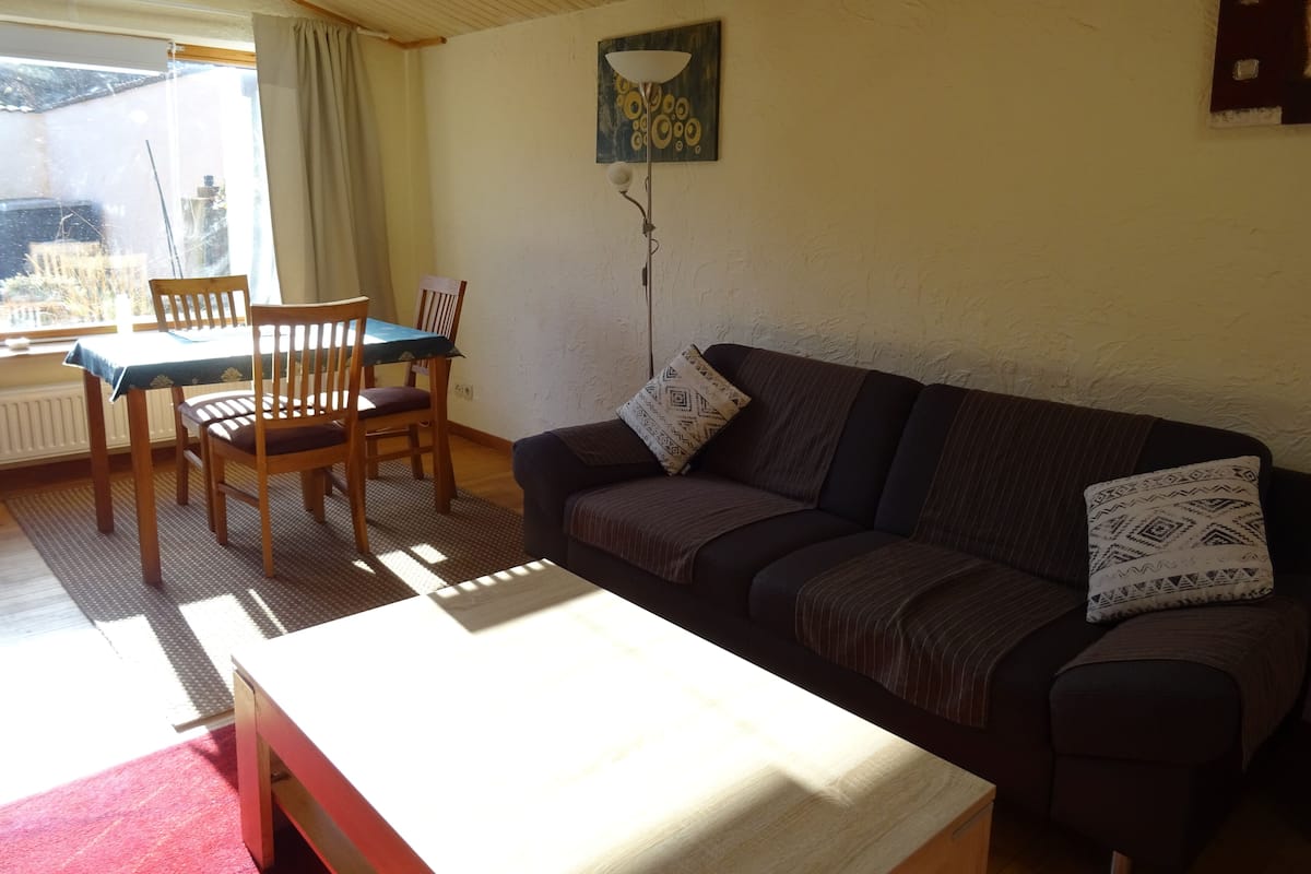 Un salon chaleureux avec une table en bois, deux chaises, un canapé agrémenté de coussins à motifs, un tableau au mur et une fenêtre avec des rideaux.