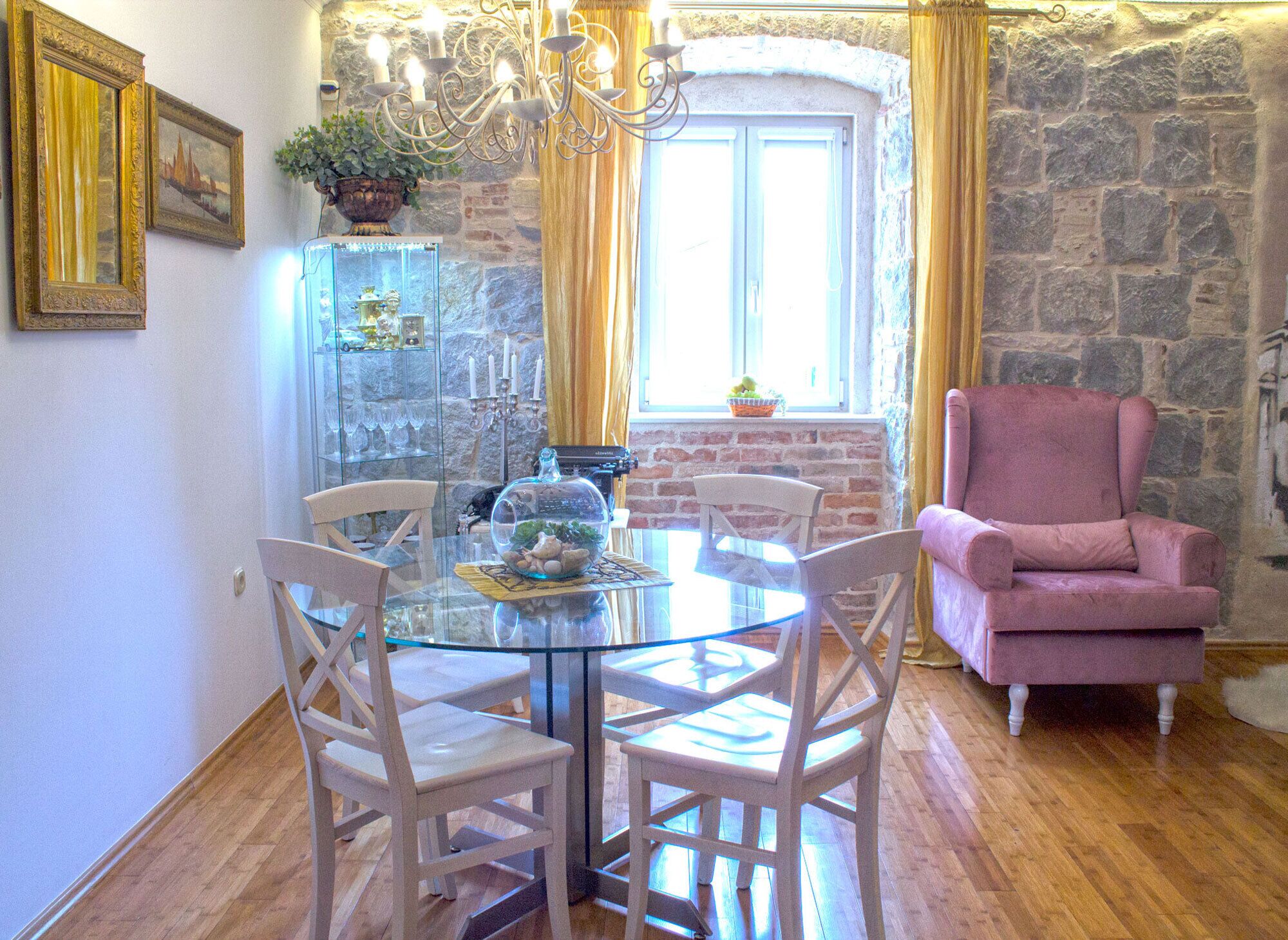 A dining area with a round glass table, white chairs, a chandelier, a stone wall, and a pink armchair.
