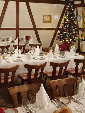 Un restaurante con varias mesas dispuestas para comer, cada una con manteles blancos, cubiertos ordenados y sillas. Se ve un árbol de Navidad decorado al fondo.