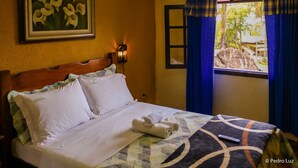 Una cama bien hecha con sábanas blancas y una toalla enrollada, un cuadro floral enmarcado en la pared y una ventana con cortinas azules.