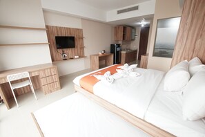 A hotel room with a bed, a desk, a chair, a TV mounted on the wall, and a kitchen area.