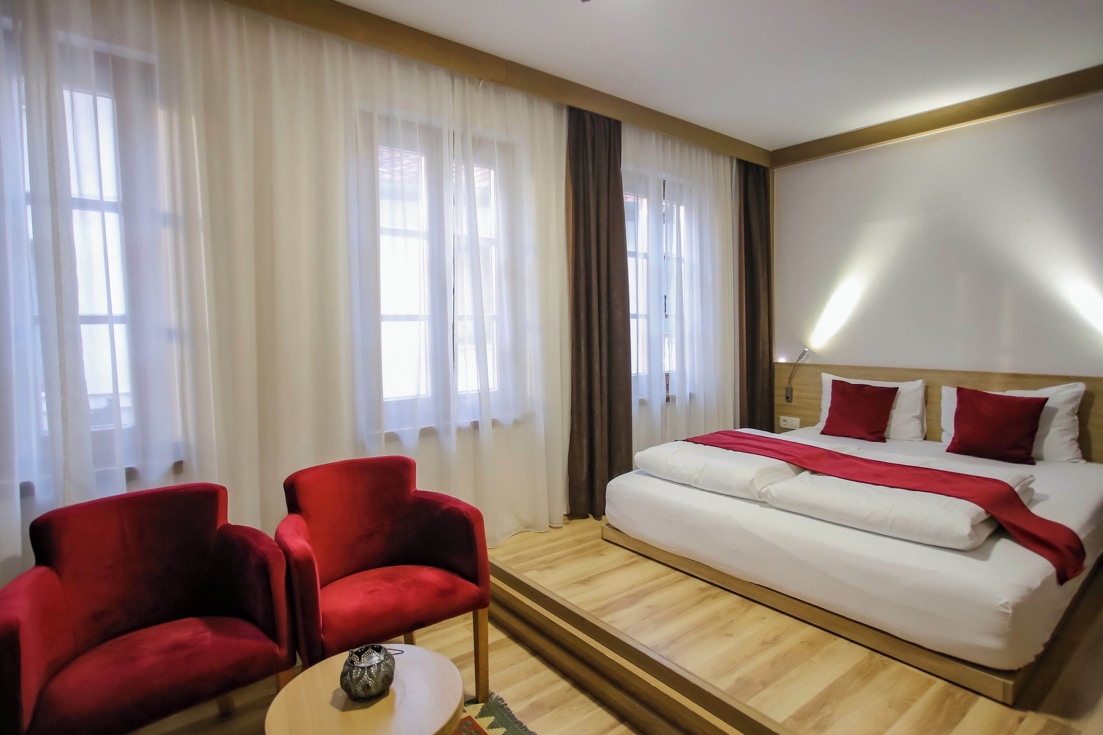 Une chambre d’hôtel avec un lit, deux fauteuils rouges, une petite table et de grandes fenêtres avec des rideaux.