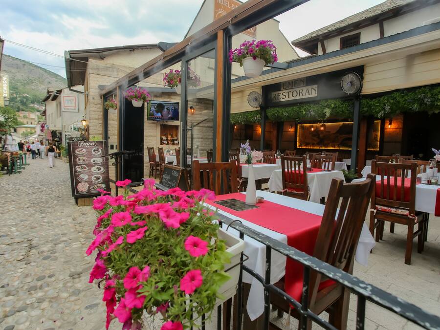 Un café al aire libre con mesas y sillas, un cartel que dice 'EMERAN RESTORAN', y una calle adoquinada.