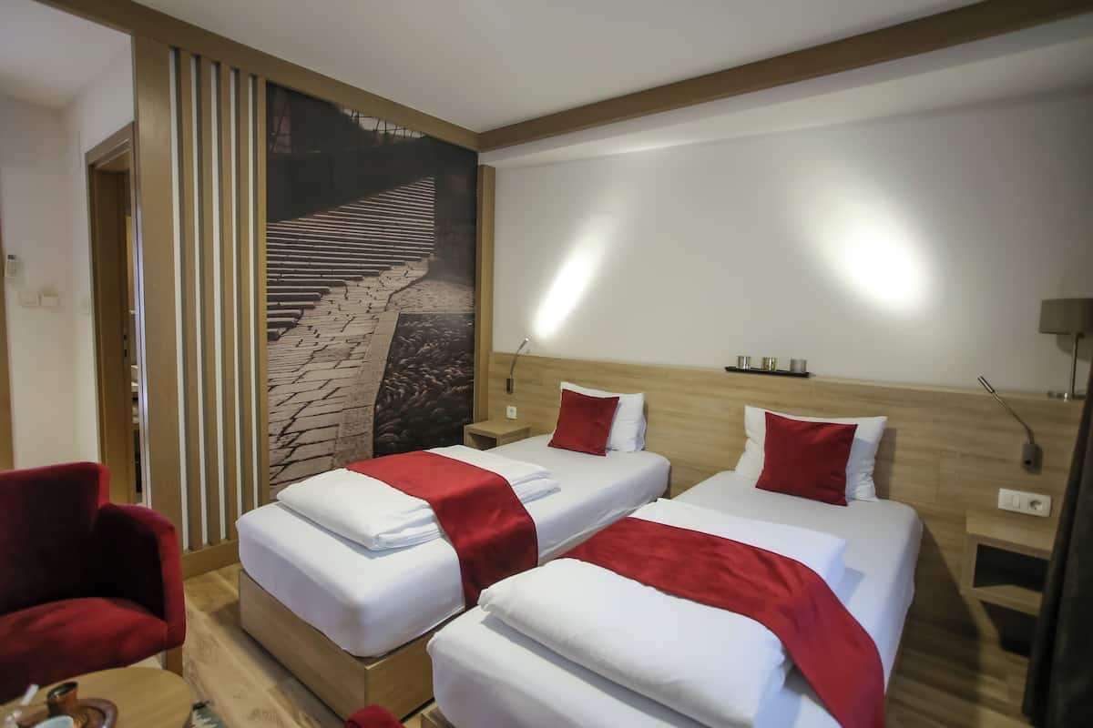 Habitación de hotel con dos camas, cada una con ropa blanca y detalles rojos, cabeceros de madera, mesitas de noche y una lámpara de pared.