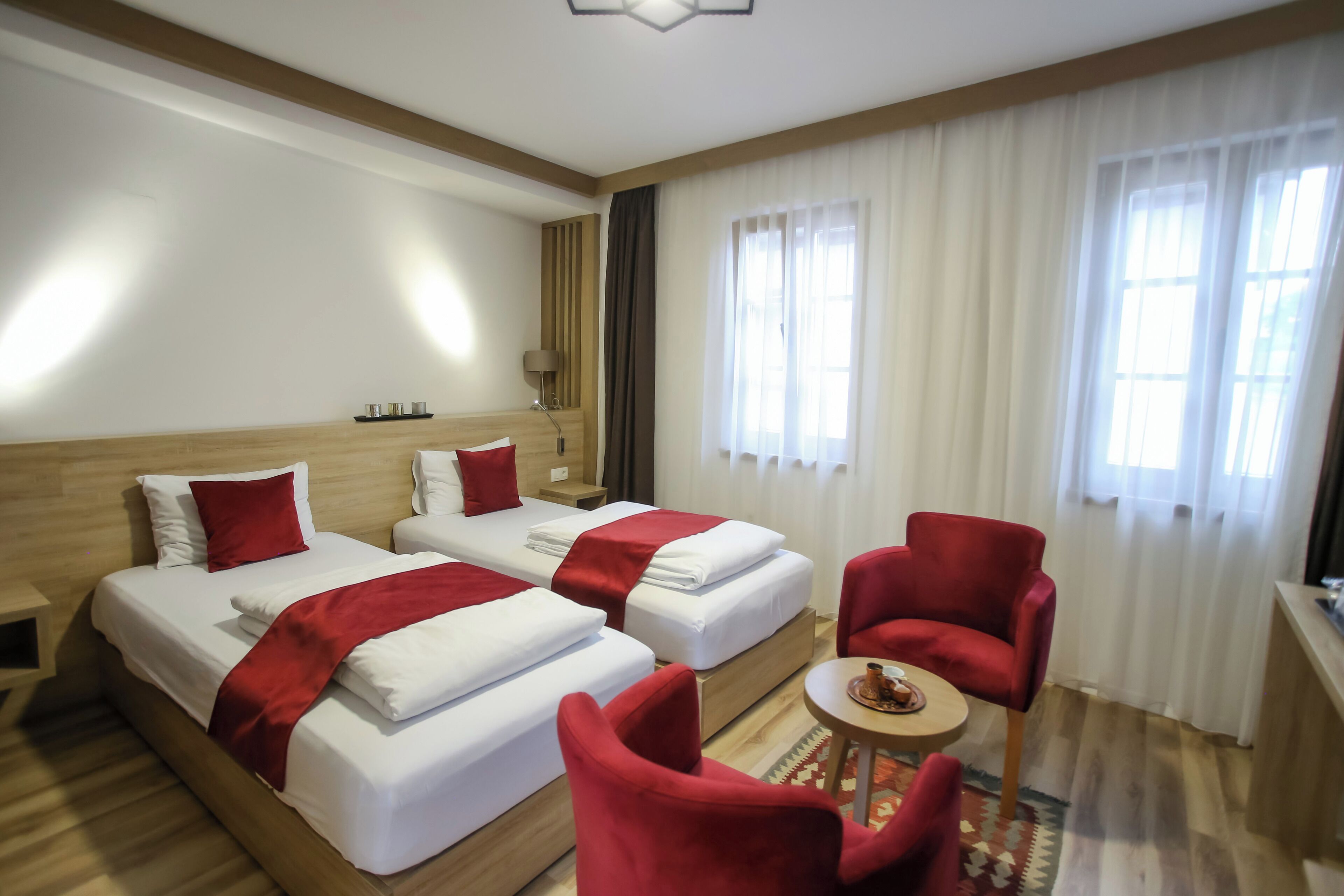 A hotel room with two beds, red chairs, a small table with a tray, and a window with curtains.