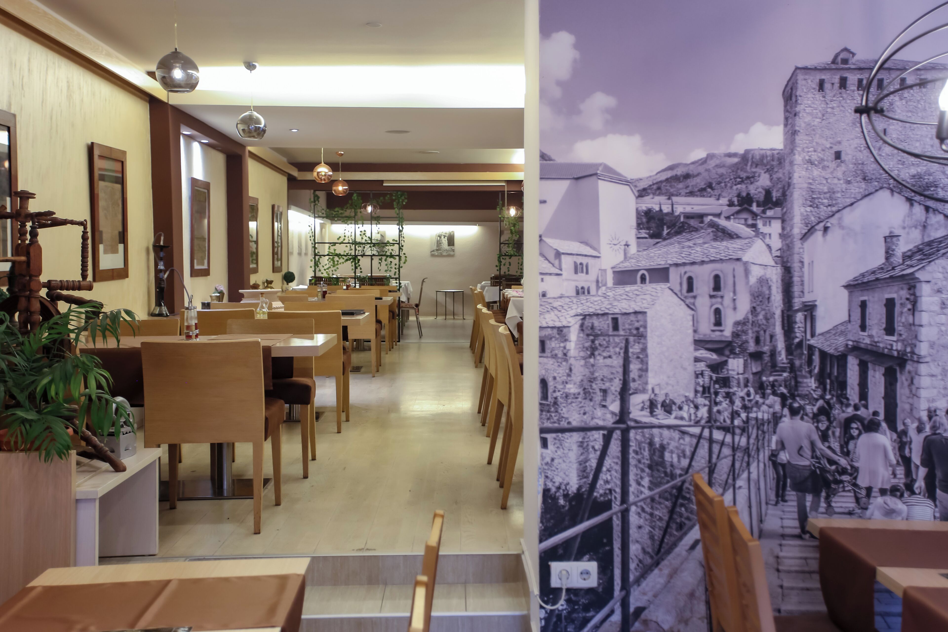 Interior de restaurante com mesas e cadeiras de madeira, plantas penduradas e um mural representando uma cena histórica de uma cidade.