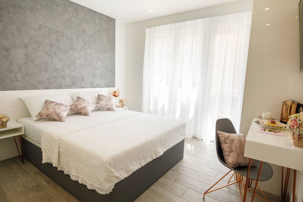 A modern bedroom with a large bed, a bedside table, a chair, and a window with sheer curtains.