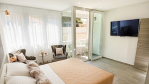 Quarto de hotel com cama grande, televisão fixada na parede, varanda com mesa e cadeiras e porta de vidro deslizante para o exterior.