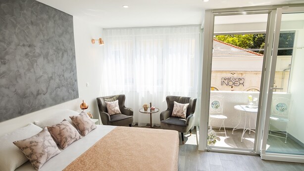 Quarto de hotel moderno com uma cama grande, duas poltronas, uma mesa pequena e uma porta de vidro deslizante que dá para uma área externa com vista para um restaurante.