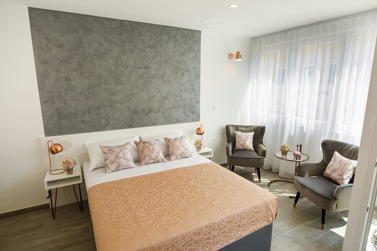 A modern hotel room with a large bed, two armchairs, a small table, and a large window with sheer curtains.