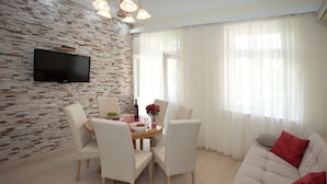 Uma sala de estar moderna com parede de pedra, TV de tela plana, mesa de jantar redonda com cadeiras e um sofá com almofadas vermelhas.