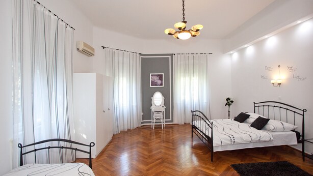 Quarto com piso em padrão herringbone de madeira, estrutura de cama em metal preto, colcha branca e tapete preto. Há uma cadeira branca, uma pequena mesa com um vaso de flores e uma luminária fixada na parede.