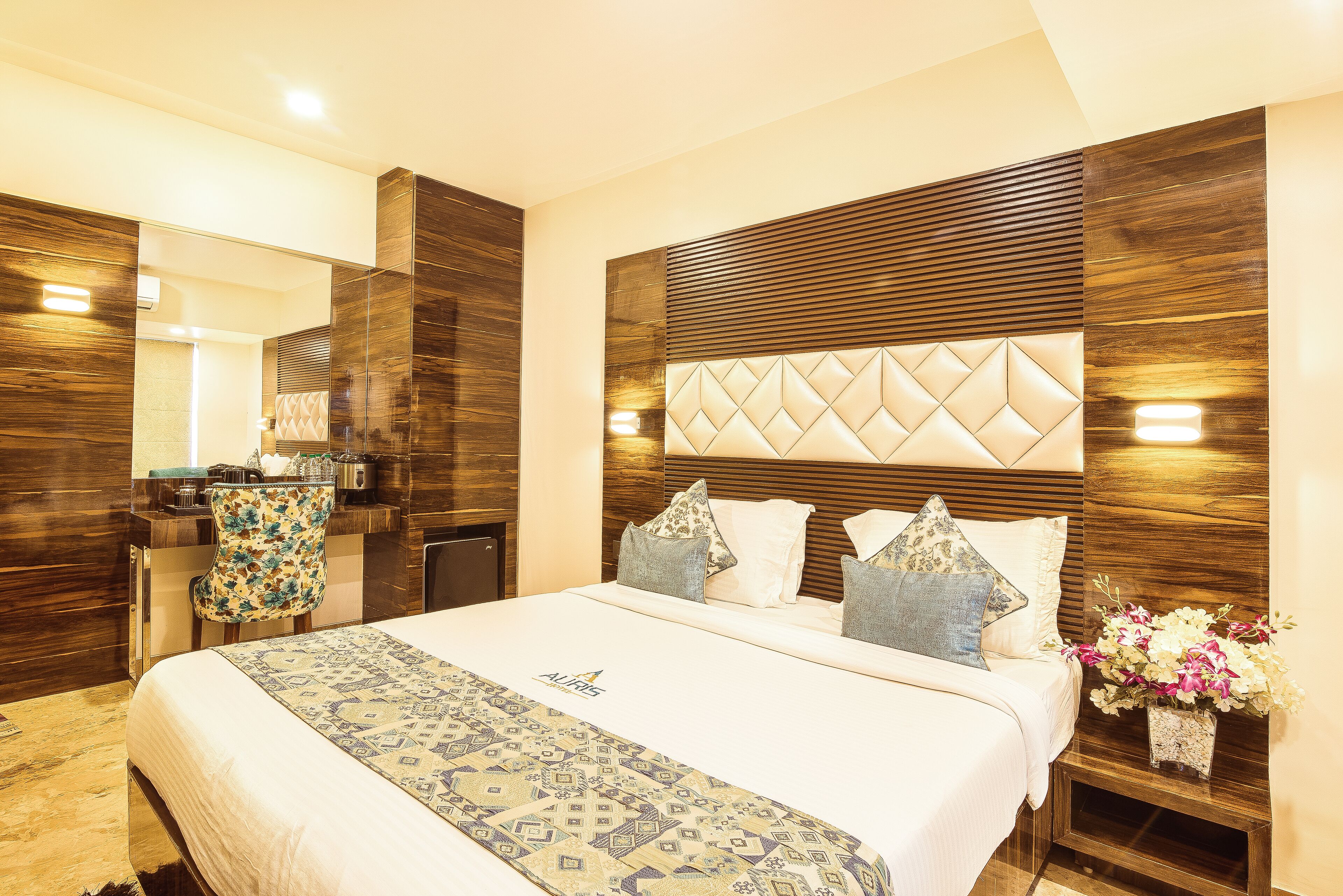 A hotel room with a large bed, a wooden headboard, a desk with a chair, and a small side table with a vase of flowers.