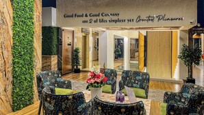 Um lobby moderno com uma parede verde, cadeiras estampadas, uma mesa com flores e uma placa com a mensagem "Good Food & Good Company".