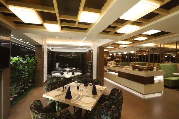A modern dining area with tables set for service, a central bar, and a seating area with green upholstered chairs.