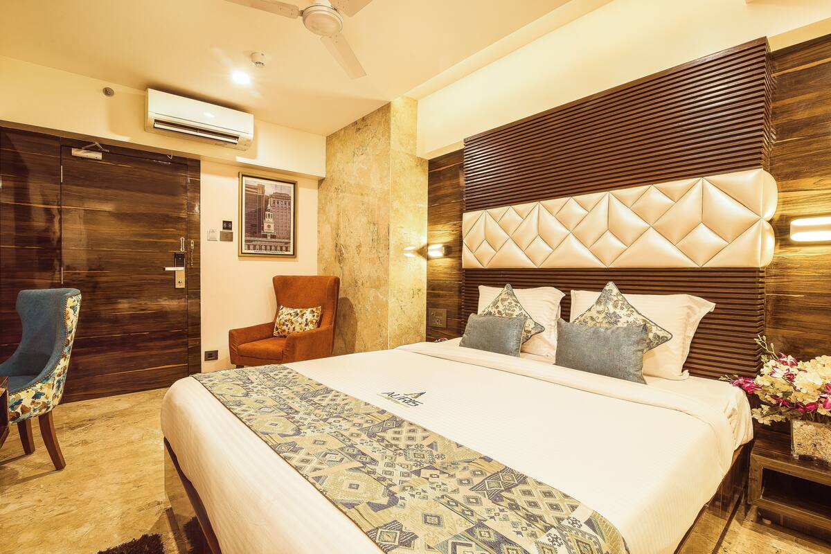 Una habitación de hotel con una cama grande, cabecera de madera, un sillón azul y un aire acondicionado instalado en la pared.