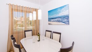 A dining area with a round table, four chairs, a bottle of wine, and a painting of a beach scene on the wall. - Cozy Villa in Okrug Gornij with swimming pool and all what is needed (Okrug Gornji)