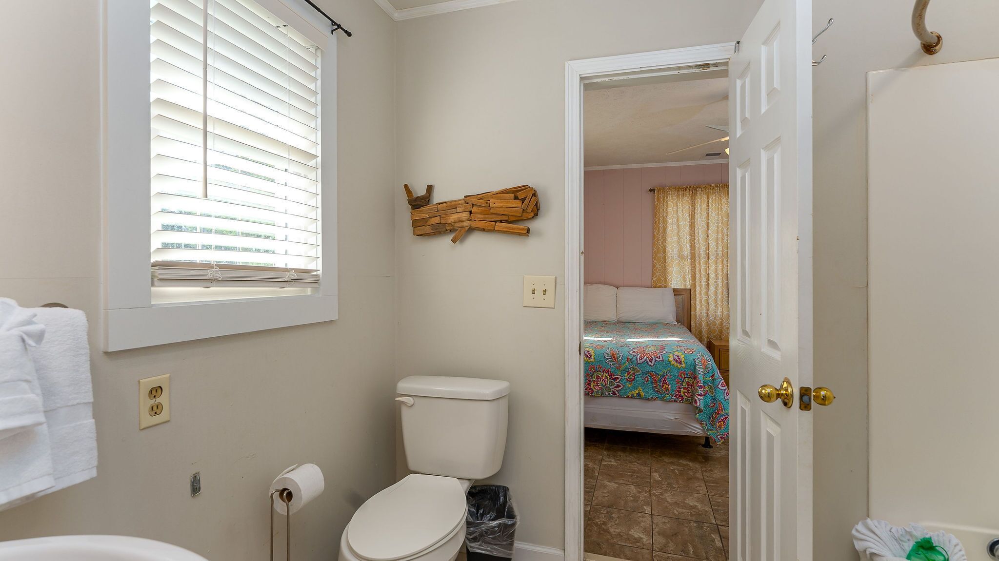 Un baño con inodoro, ventana con persianas, adorno de madera en la pared, y puerta que da a un dormitorio con cama y cortinas.