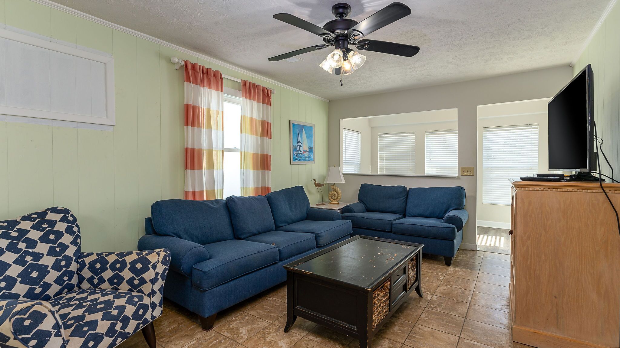 A living room with two blue sofas, a patterned armchair, a wooden coffee table, a ceiling fan, and a television on a wooden cabinet.