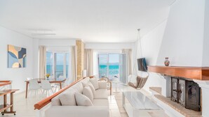 Een moderne woonkamer met een bank, salontafel en televisie, met een open haard en uitzicht op zee.