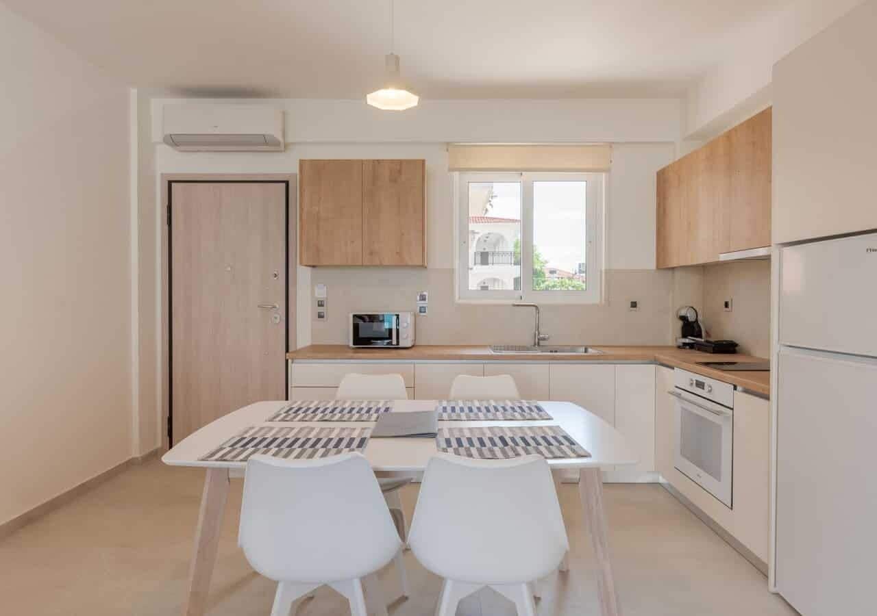 Una cocina moderna con armarios blancos, microondas integrado y refrigerador. Hay una mesa de comedor con dos sillas y un ventanal que proporciona luz natural.