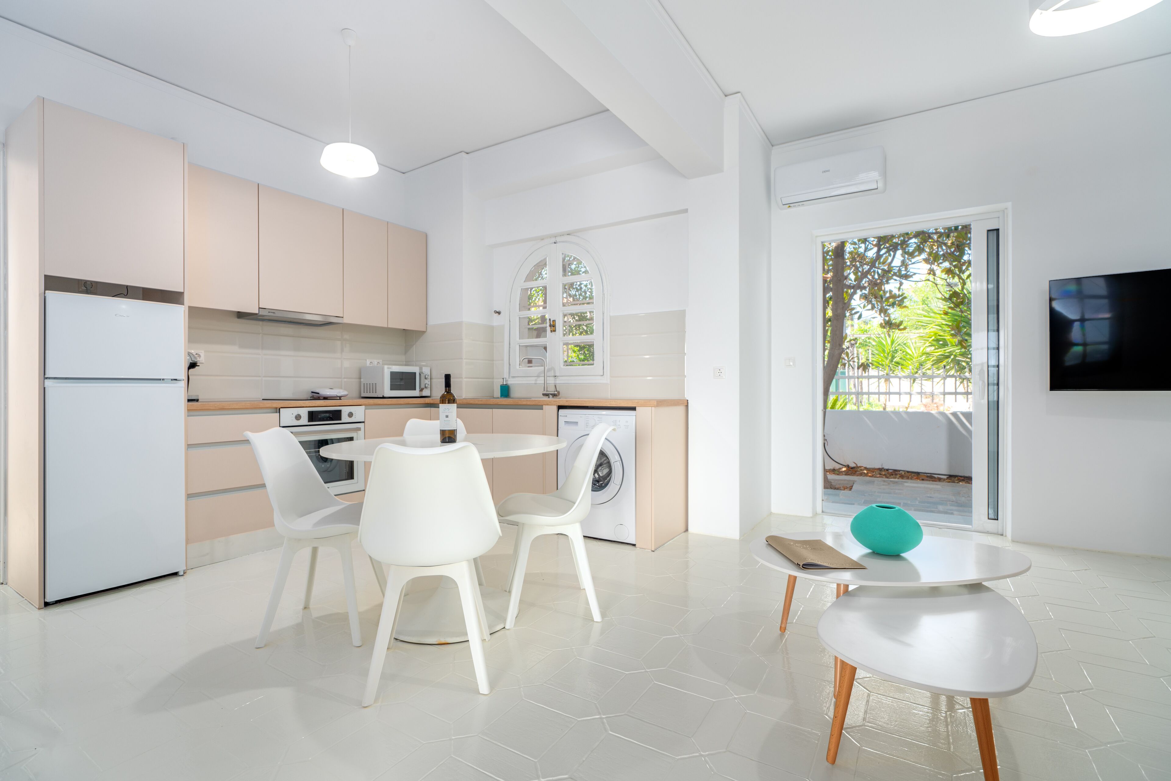 A modern kitchen with white appliances, a dining area with white chairs, and a view of greenery outside.