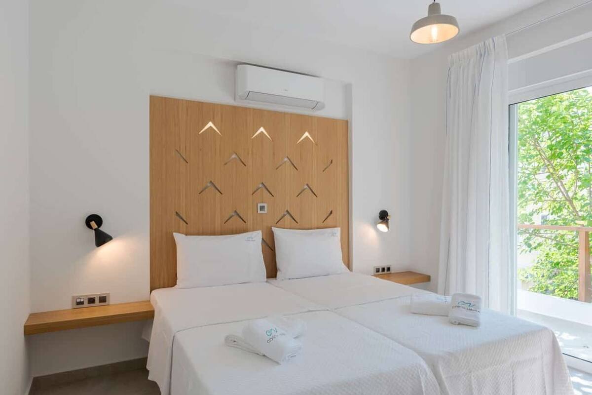 Habitación de hotel con cabecera de madera, dos camas, mesitas de noche, ventana con cortinas y vista a zonas verdes.