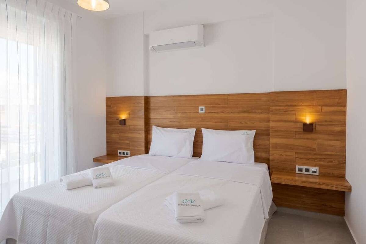 Habitación de hotel con dos camas, ropa de cama blanca, cabecero de madera, lámparas de noche y una ventana con cortinas.